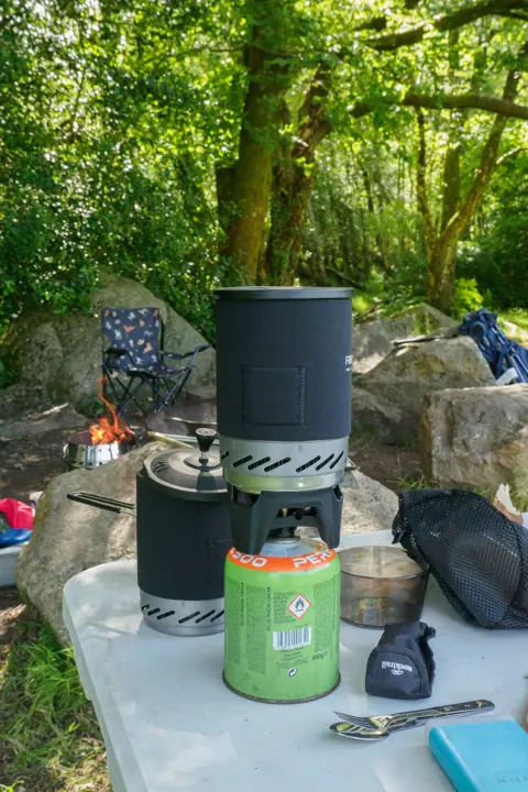 A camp kitchen on a table by a river, with a gas stove, kettle and pots, and a small fire and folding chair behind.