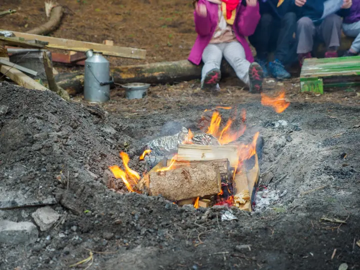 A campfire burns in a pit, with children in coloured jackets sitting on logs in the background.