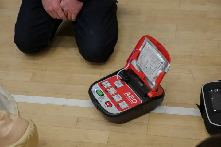 An AED defibrillator open on a wooden hall floor next to a kneeling person and a training manikin.