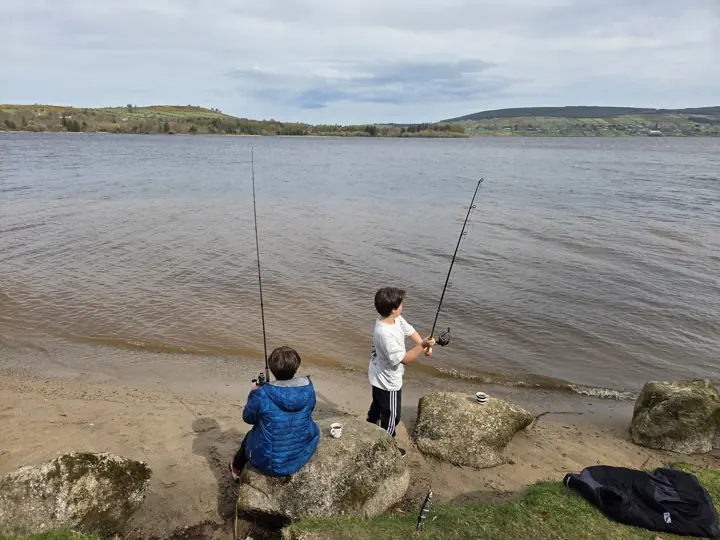 Two children fishing from a sandy lakeshore, one sitting on a rock and one standing, both seen from behind.