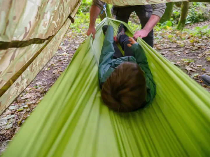 A child lies in a green hammock under a tarp while an adult holds the edge.