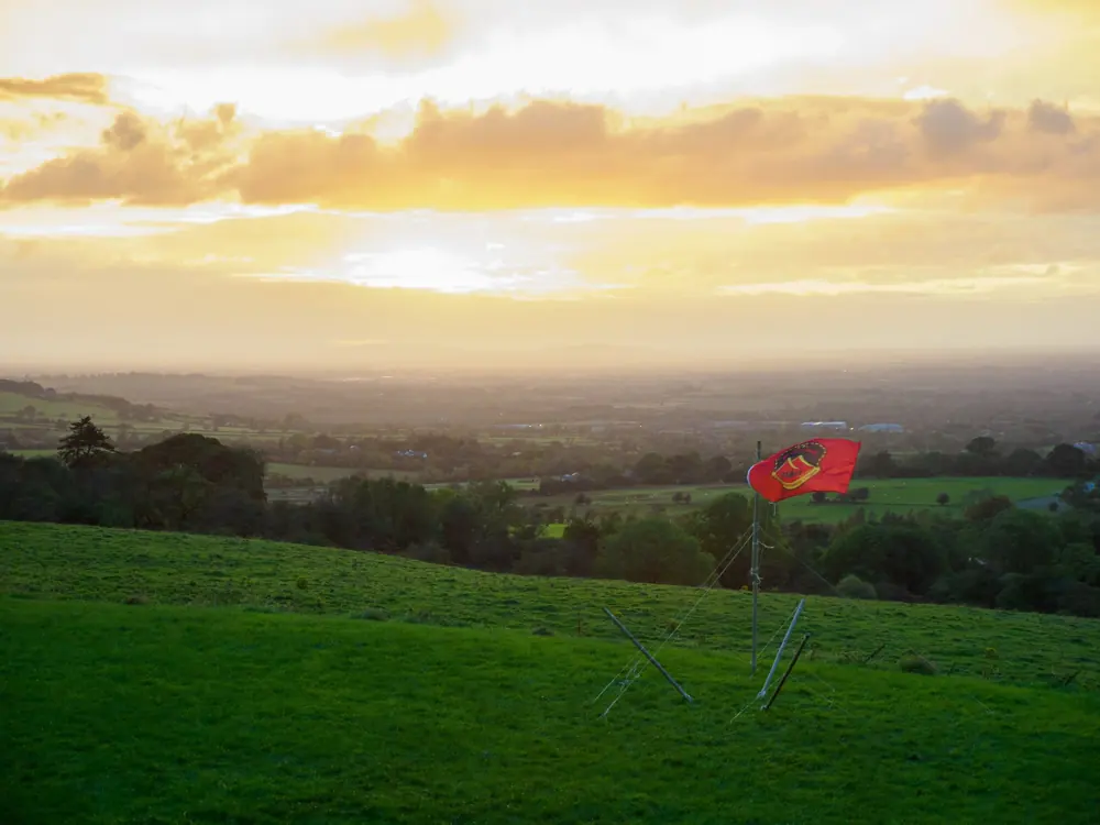 A red group flag flies on a hilltop campsite at sunset, with green fields and a golden sky beyond.