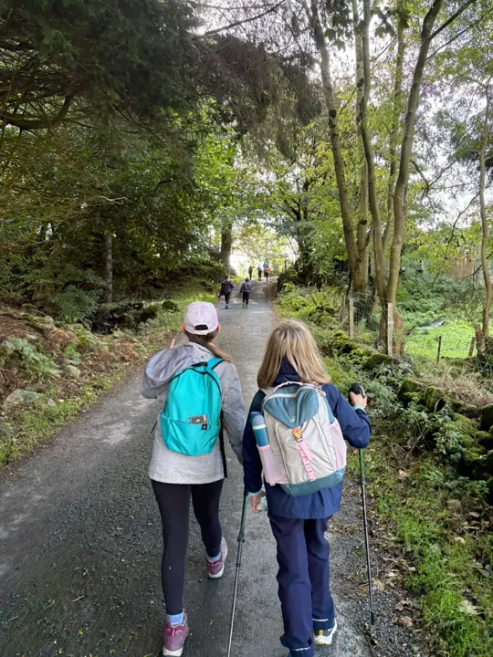 Two children with backpacks and walking poles walk along a tarmac path through woodland, seen from behind.