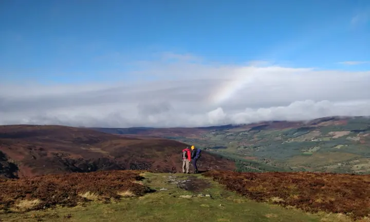 Two hikers in red and navy jackets with backpacks stand on a heather-covered hilltop looking over a valley, with a cloud bank and a faint rainbow behind them.