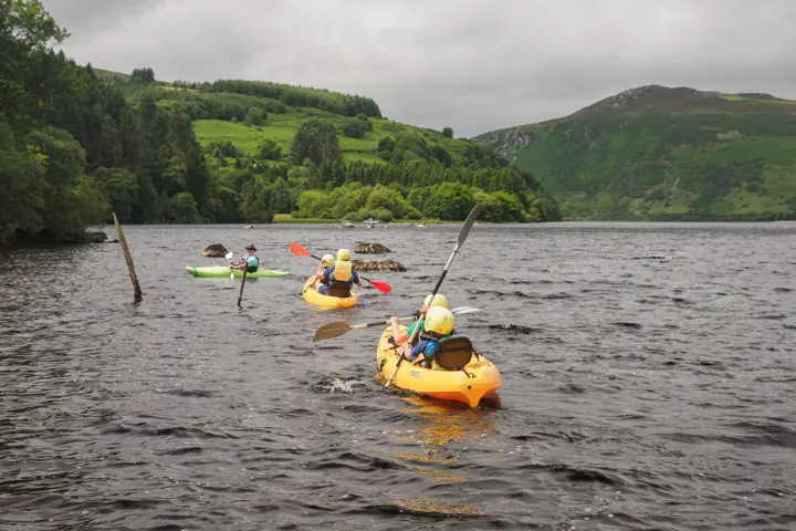 Children in yellow kayaks paddle across a lake, seen from behind, with green hills beyond.