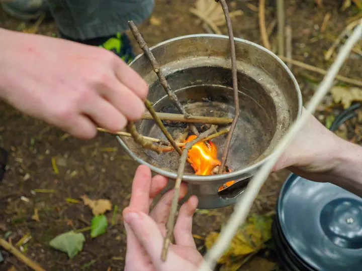 Hands feed small twigs into a metal pan to light a small fire.