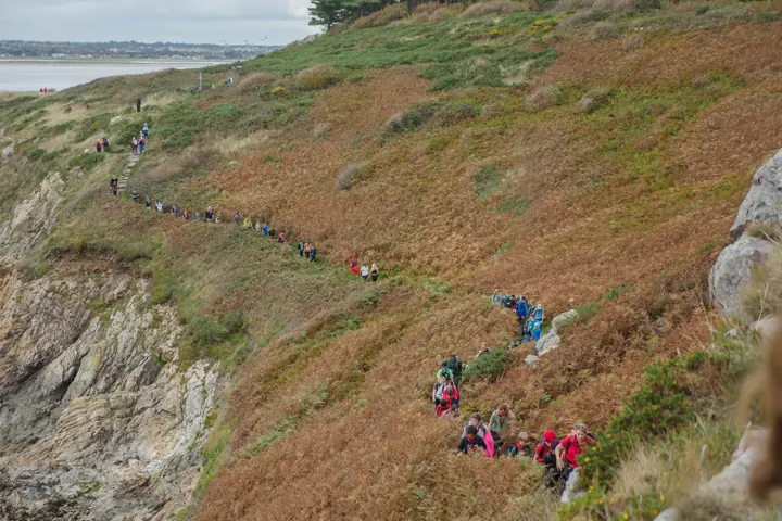 A long line of walkers follows a coastal path along a bracken-covered hillside above the sea.
