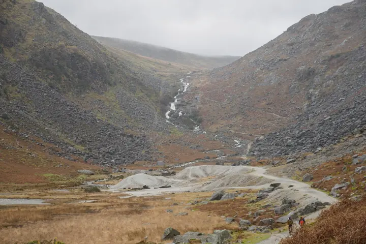 A rocky mountain valley with a stream running down to a flat gravel area, under grey cloud.
