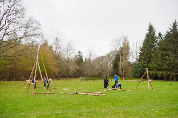 Scouts working on wooden pioneering frames in a grassy field with trees behind.