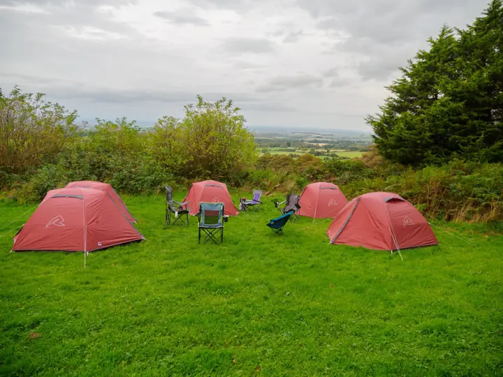 Red tents and camping chairs on a bright green hilltop pitch with countryside beyond.