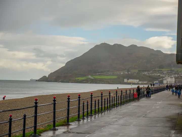 A seafront promenade with railings and a headland across the bay, with walkers in the distance.