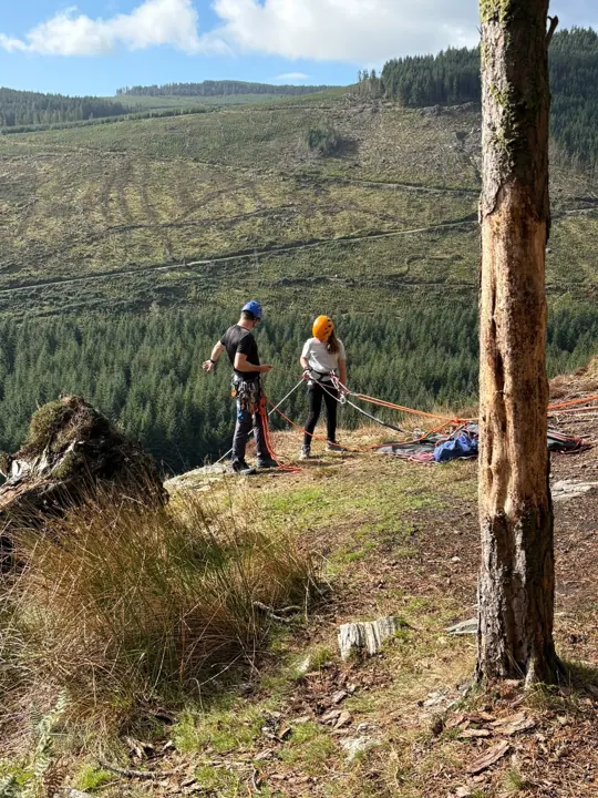 Two leaders in helmets set up ropes beside a tree on a forested hillside.