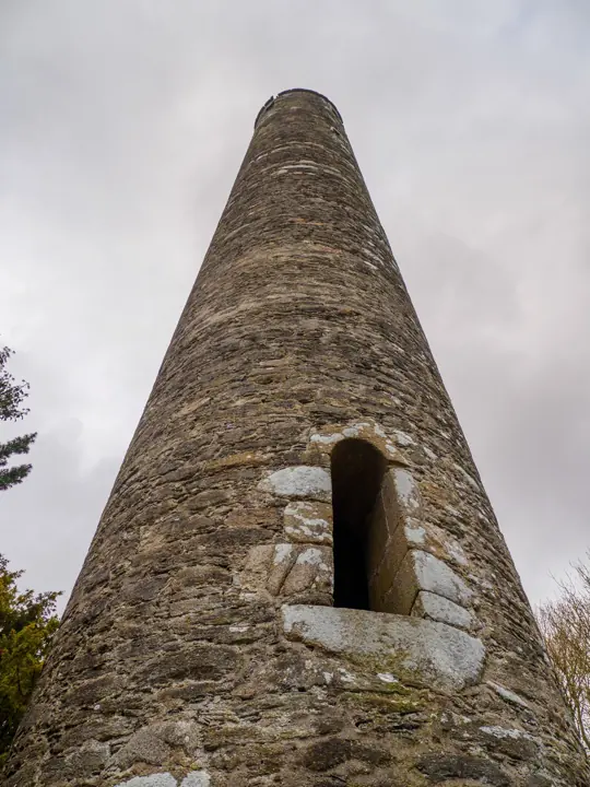 Looking up at a tall stone round tower with a small doorway, against a grey sky.