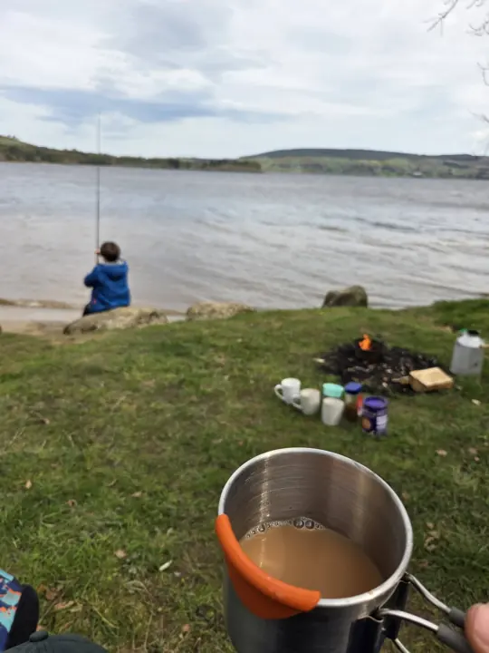 A child in a blue jacket fishes from a grassy lakeshore, with mugs and a small campfire on the grass and a steel mug of tea in the foreground.