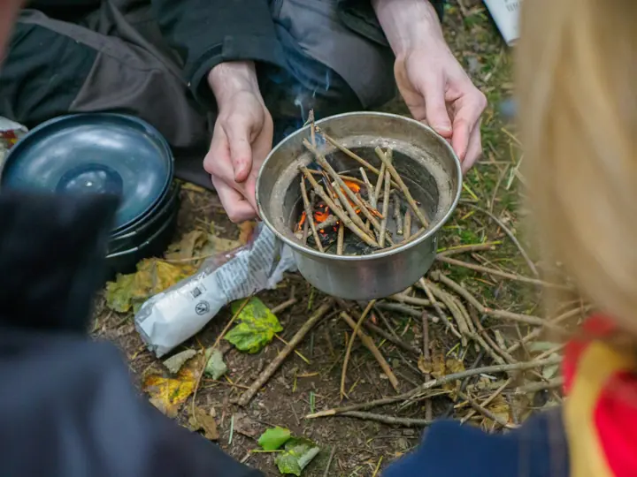 Hands tend a small twig fire in a metal bowl, with a child in a red neckerchief at the edge of the picture.