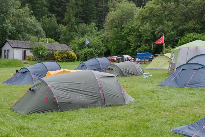 Dome tents pitched on a campsite with a flag flying behind, near a small building and trees.
