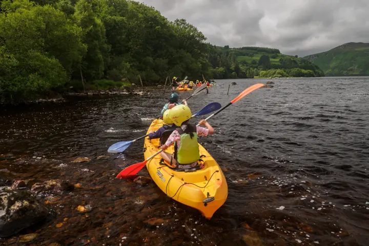 Two children in a yellow kayak with orange, red and blue paddles, seen from behind on dark lake water.